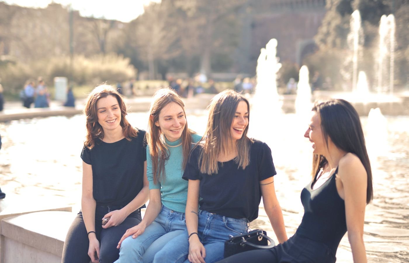 Four female friends laughing together by a fountain on a sunny day